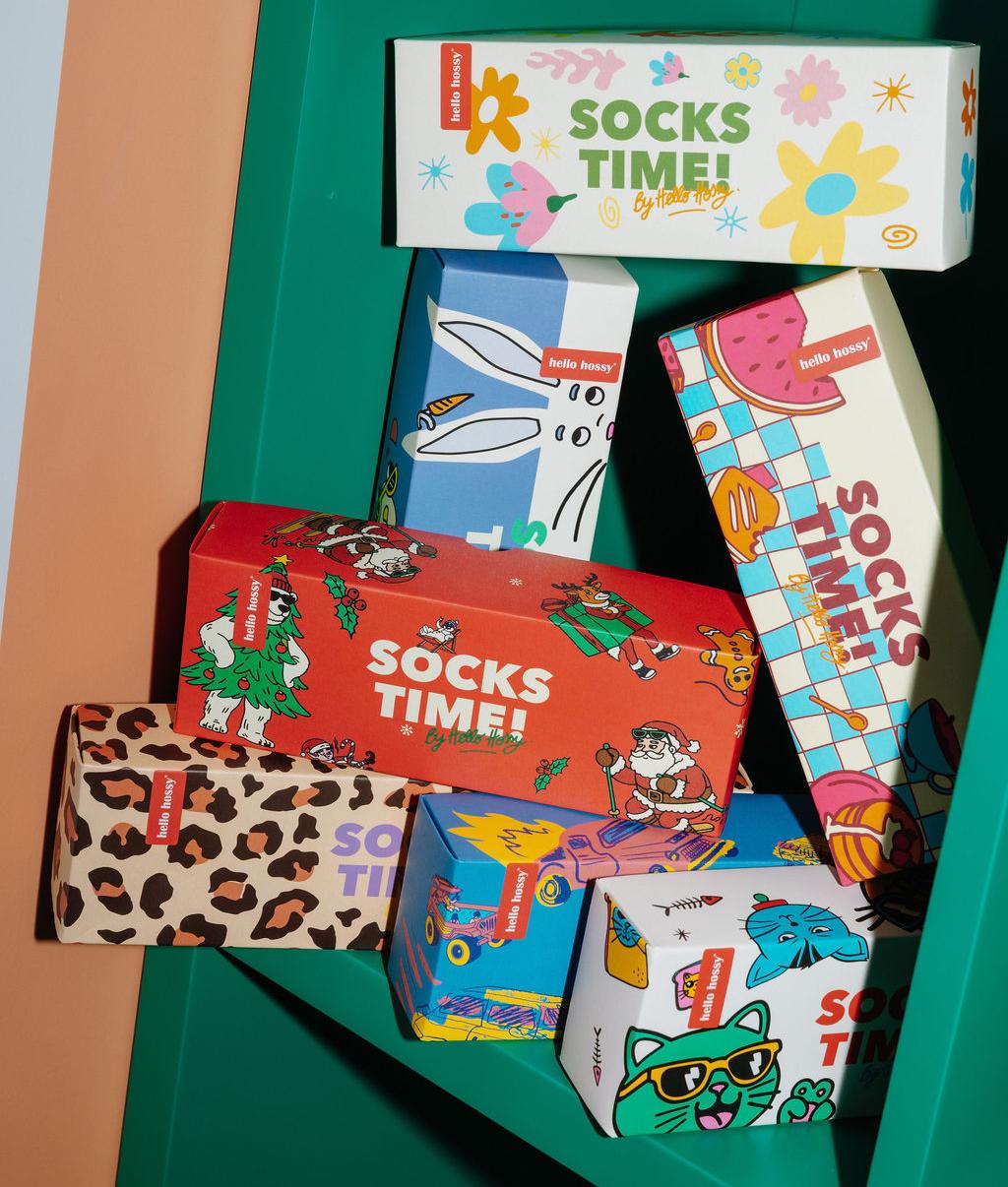 Coffrets cadeaux "Socks Time !" empilés sur une étagère verte, packaging colorés et illustrés : motifs Noël, fleurs, léopard, chats, mascottes, voiture et nourriture - Hello Hossy